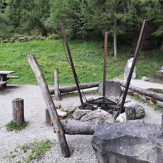 A cozy outdoor grill area with a large fire circle made of stones and wood. In the background, there is a picnic bench on a green meadow. | © Kleinwalsertal Tourismus | Veronika Senn