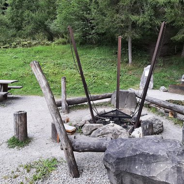 A cozy outdoor grill area with a large fire circle made of stones and wood. In the background, there is a picnic bench on a green meadow. | © Kleinwalsertal Tourismus | Veronika Senn