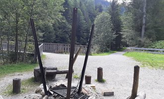 A fire pit in a forested area, surrounded by trees and a gravel path. In the background, logs and a bridge are visible. | © Kleinwalsertal Tourismus | Veronika Senn