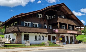 A charming, traditional wooden chalet with multiple balconies and windows. The sky is blue and filled with clouds, while the green lawn surrounds the building. | © Antik und Kunst im Malerhäusl Herbert Stumvoll