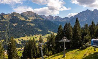 Eine malerische Berglandschaft mit grünen Wiesen und hohen Tannenbäumen. Im Hintergrund sind majestätische Berge und ein Skilift zu sehen. | © OBERSTDORF · KLEINWALSERTAL BERGBAHNEN