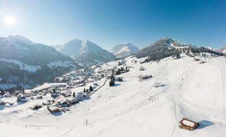 Eine malerische Winterlandschaft mit schneebedeckten Bergen und klar blauem Himmel. Im Vordergrund sind Skipisten und ein kleines Dorf in der Ferne zu sehen. | © OBERSTDORF · KLEINWALSERTAL BERGBAHNEN