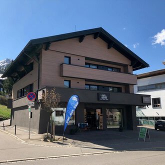 A modern building at a street corner with a shop on the ground floor. In the background, mountains and other buildings can be seen. | © Sport Hilbrand OG