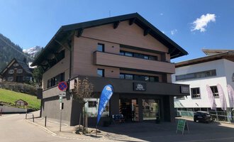 A modern building at a street corner with a shop on the ground floor. In the background, mountains and other buildings can be seen. | © Sport Hilbrand OG