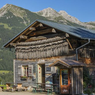 A traditional wooden house in the Alps with a green landscape background. The mountains are clearly and sunnily visible. | © Hoflaada | Dagmar Hilbrand