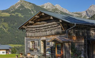 A traditional wooden house in the Alps with a green landscape background. The mountains are clearly and sunnily visible. | © Hoflaada | Dagmar Hilbrand