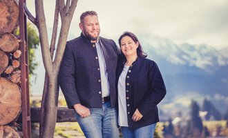 A couple stands smiling in front of a picturesque mountain landscape. Both are dressed in traditional clothing and appear friendly and relaxed. | © Alpenhof Jäger | Werbewind