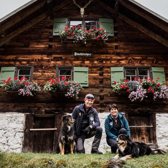 Ein rustikales Holzhaus mit blühenden Blumenkästen. Ein Paar mit zwei Hunden posiert davor. | © Hundewelt Kleinwalsertal | Stefan Lindbauer