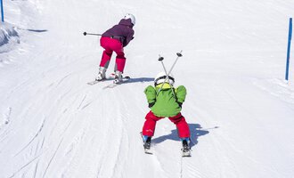 Zwei Kinder fahren Ski auf einer verschneiten Piste. Das eine Kind trägt einen grünen Anzug und das andere einen lila Anzug. | © Kleinwalsertal Tourismus | Dominik Berchtold