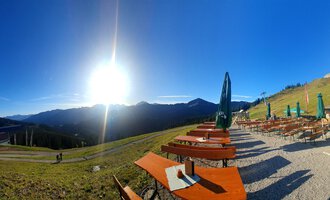 A beautiful mountain landscape with the sun shining over the peaks. In the foreground, there are wooden benches and tables with green sunshades. | © Ifenhütte | Petra Diener