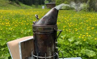A smoking device is on a table in a blooming meadow. In the background, forests and a blue sky can be seen. | © Imkerei Jürgen Scharnagl