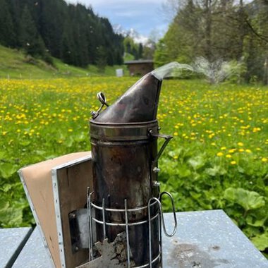 Ein Rauchgerät steht auf einem Tisch in einer blühenden Wiese. Im Hintergrund sind Wälder und ein blauer Himmel zu sehen. | © Imkerei Jürgen Scharnagl