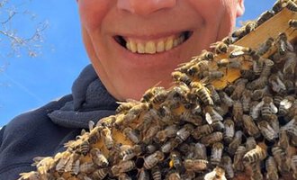 A man smiles in front of a beehive covered with many bees. The sky is blue and sunny. | © Imkerei Jürgen Scharnagl
