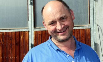 Ein fröhlicher Mann lächelt in die Kamera. Im Hintergrund sind Holztüren und Fenster zu sehen. | © Innere Wiesalpe | Andrea Kainz zuckerschnecke.at