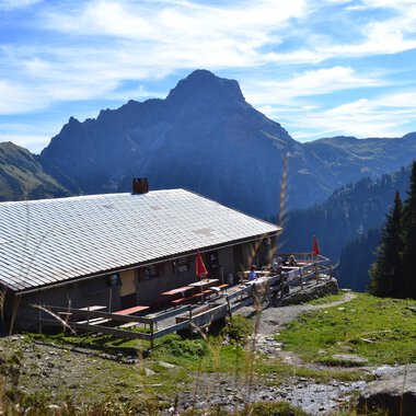 Eine alpine Hütte umgeben von Bergen und Wäldern. Der Himmel ist klar mit ein paar Wolken. | © Kleinwalsertal Tourismus