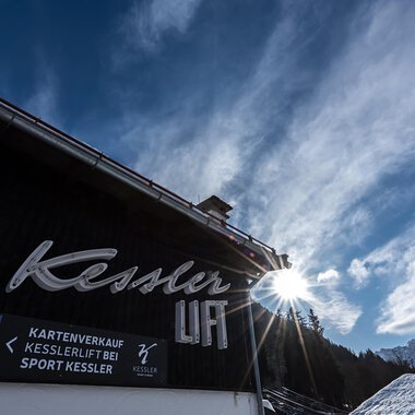 A building with the inscription "Kessler" and a sunburst in the background. The sky is blue with some clouds and the surroundings look alpine. | © Sport Kessler GmbH