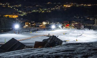 A snowy ski slope at night with illuminated lifts. In the background, the lights of a small village are visible. | © Sport Kessler GmbH