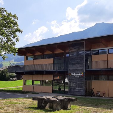 Ein modernes Gebäude mit großen Fenstern und Holzverkleidung. Im Vordergrund steht ein Steintisch auf einer grünen Wiese mit Bergen im Hintergrund. | © Kleinwalsertal Tourismus | Veronika Senn
