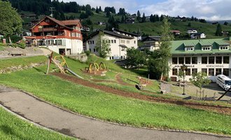 A picturesque landscape with green meadows and playgrounds. In the background, some chalet-like buildings and mountains can be seen. | © Kleinwalsertal Tourismus |