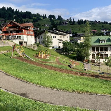 A picturesque landscape with green meadows and playgrounds. In the background, some chalet-like buildings and mountains can be seen. | © Kleinwalsertal Tourismus |
