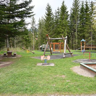 A playground in a forest with several play devices and a lawn. In the background, there are tall trees and a picnic bench. | © Gemeinde Mittelberg