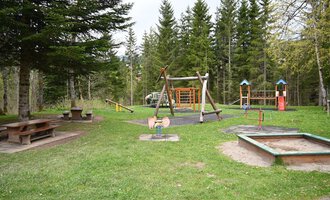 A playground in a forest with several play devices and a lawn. In the background, there are tall trees and a picnic bench. | © Gemeinde Mittelberg