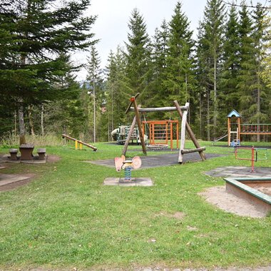 A playground in a forest with several play devices and a lawn. In the background, there are tall trees and a picnic bench. | © Gemeinde Mittelberg