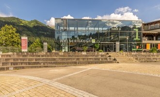 A modern building with a glass facade and the inscription "WALSERHAUS". In the background, green mountains and a blue sky are visible. | © Kleinwalsertal Tourismus | Steffen Berschin