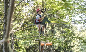 A boy is sitting on a rope in a high ropes course. The surroundings are surrounded by trees, and he is wearing a helmet and safety gear. | © OBERSTDORF · KLEINWALSERTAL BERGBAHNEN