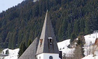 Eine malerische Kirche mit einem spitzen Turm steht im Schnee. Im Hintergrund erheben sich bewaldete Berge. | © Ev. Kirche Kleinwalsertal