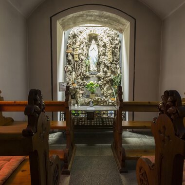 A quiet chapel with wooden benches and a statue in the niche. The walls are softly illuminated, creating a peaceful atmosphere. | © Kleinwalsertal Tourismus | Oliver Farys