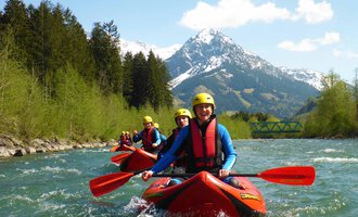 A group of kayakers is paddling on a clear river. In the background, there are snow-capped mountains and green trees. | © MAP-Erlebnis