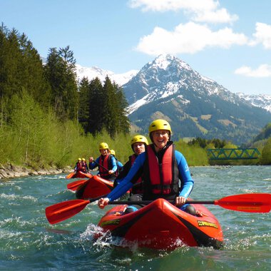 A group of kayakers is paddling on a clear river. In the background, there are snow-capped mountains and green trees. | © MAP-Erlebnis