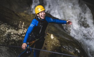 A person in a blue wetsuit and yellow helmet stands in a rocky environment. They are smiling and appear happy, while water flows in the background. | © MAP-Erlebnis