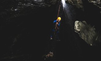 A climber hangs on a rope in a dark gorge. Water cascades down, creating an exhilarating atmosphere. | © MAP-Erlebnis