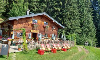 A cozy wooden house surrounded by tall trees. In front of the house, there are chairs and tables in an inviting outdoor area. | © Maxhütte | Viera Krafcik