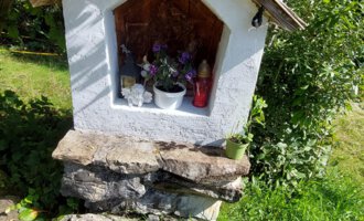 A small white chapel with a gable roof stands on a stone foundation. Surrounded by green grass and trees, it is decorated with flowers. | © Maxhütte | Viera Krafcik