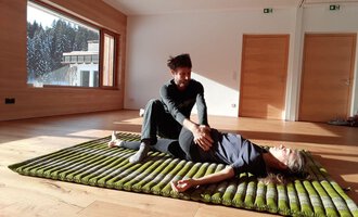 A person is lying on a mat while another person gently treats them. The room has large windows overlooking trees and plenty of daylight. | © Alexander Mayr