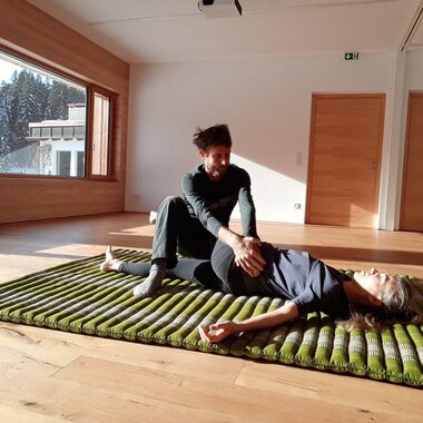 A person is lying on a mat while another person gently treats them. The room has large windows overlooking trees and plenty of daylight. | © Alexander Mayr