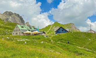 Eine malerische Berglandschaft mit gemütlichen Hütten und grünen Wiesen. Im Hintergrund sind beeindruckende Felsen und ein blauer Himmel mit Wolken zu sehen. | © Mindelheimer Hütte | Wolfgang Stache