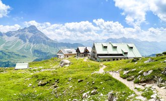 Eine malerische Berglandschaft mit grünem Gras und einem klaren Himmel. Im Vordergrund sind typische Berghütten zu sehen. | © Mindelheimer Hütte | Wolfgang Stache