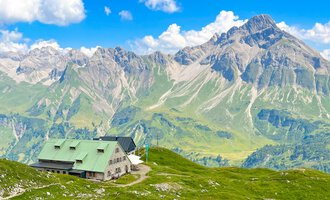 Eine Berghütte liegt malerisch auf einer grünen Wiese. Im Hintergrund erheben sich beeindruckende Berge unter einem blauen Himmel mit einigen Wolken. | © Mindelheimer Hütte | Wolfgang Stache
