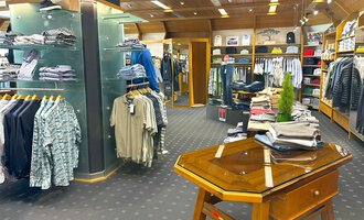 Ein moderner Bekleidungsladen mit verschiedenen Shirts und Hosen. Im Vordergrund steht ein Holztisch, während die Regale im Hintergrund voll mit Kleidung sind. | © Moosbrugger Mode e.U.