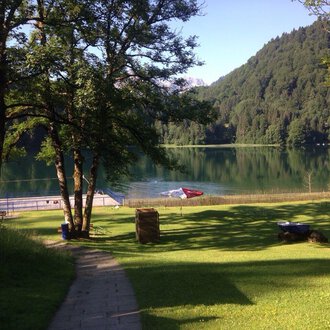 A tranquil lake surrounded by trees and green meadows. In the background, mountains and sun loungers by the shore can be seen. | © Kleinwalsertal Tourismus | Naturbad Freibergsee