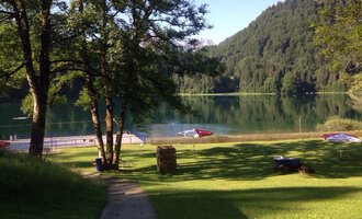 A tranquil lake surrounded by trees and green meadows. In the background, mountains and sun loungers by the shore can be seen. | © Kleinwalsertal Tourismus | Naturbad Freibergsee