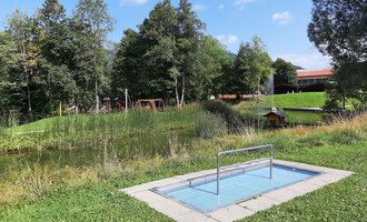 Ein kleiner Swimmingpool in einer grünen Wiese, umgeben von Bäumen. Im Hintergrund sind Spielgeräte und ein Gebäude zu sehen. | © Kleinwalsertal Tourismus | Veronika Senn