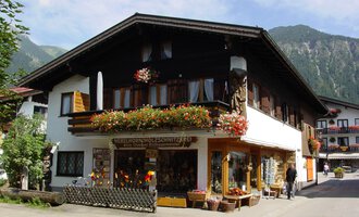 A traditional house in the mountains, surrounded by greenery. The shop on the ground floor offers handcrafted wooden carvings and is adorned with flowers. | © Nebelhorn Holzschnitzerei e.K.