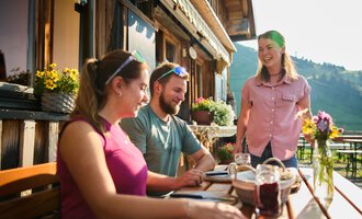 A friendly outdoor atmosphere with two guests at a table. A server is serving the food in a nice, rustic establishment. | © Kleinwalsertal Tourismus | Oliver Farys