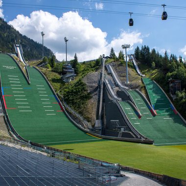 Zwei große Sprungschanzen umgeben von grünen Wiesen und Bergen. Im Hintergrund hängen Seilbahnen unter einem blauen Himmel. | © Skiclub Oberstdorf Veranstaltungs GmbH