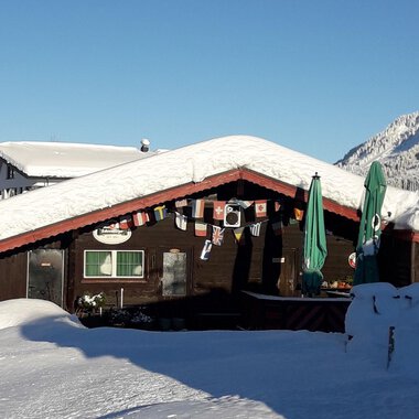 Eine gemütliche Hütte im Schnee, umgeben von einer winterlichen Landschaft. Der Himmel ist klar und die Berge sind gut sichtbar.
